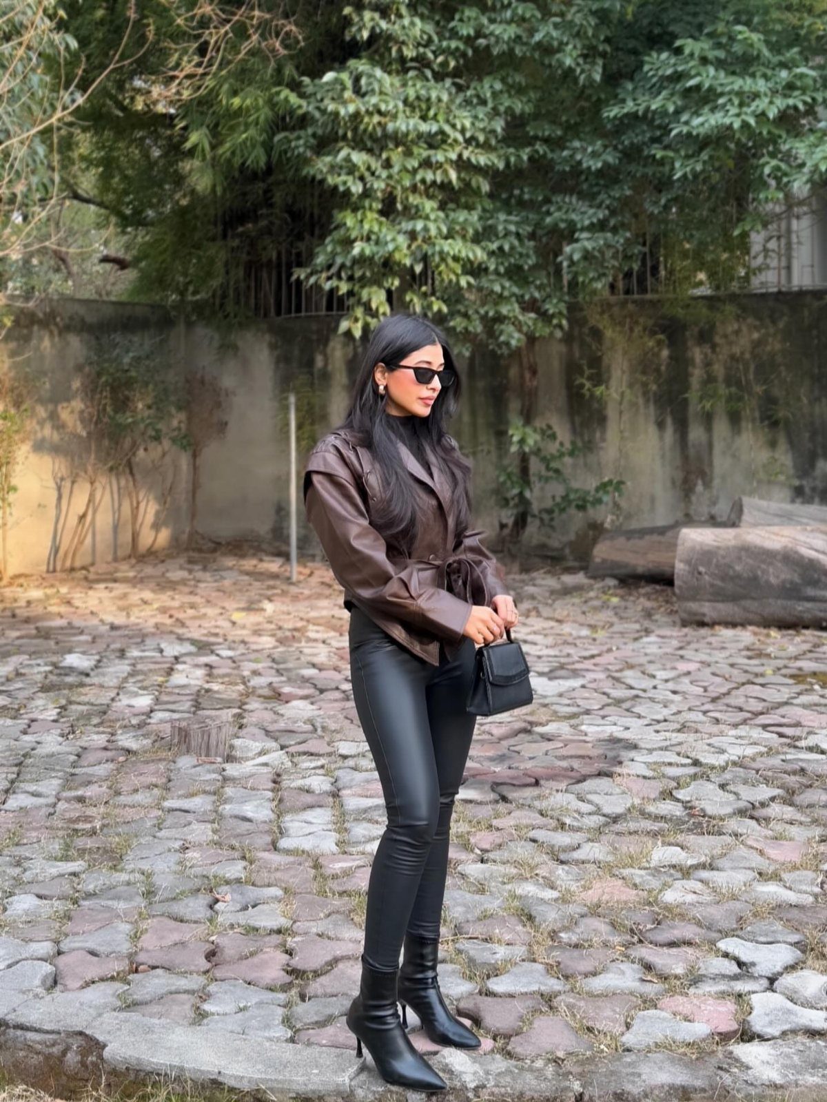 Woman wearing a brown leather jacket, black faux leather pants, and black boots standing outdoors.