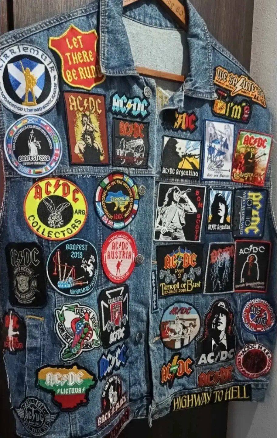 A blue denim jacket covered in various colorful AC/DC band patches hanging on a wooden hanger.