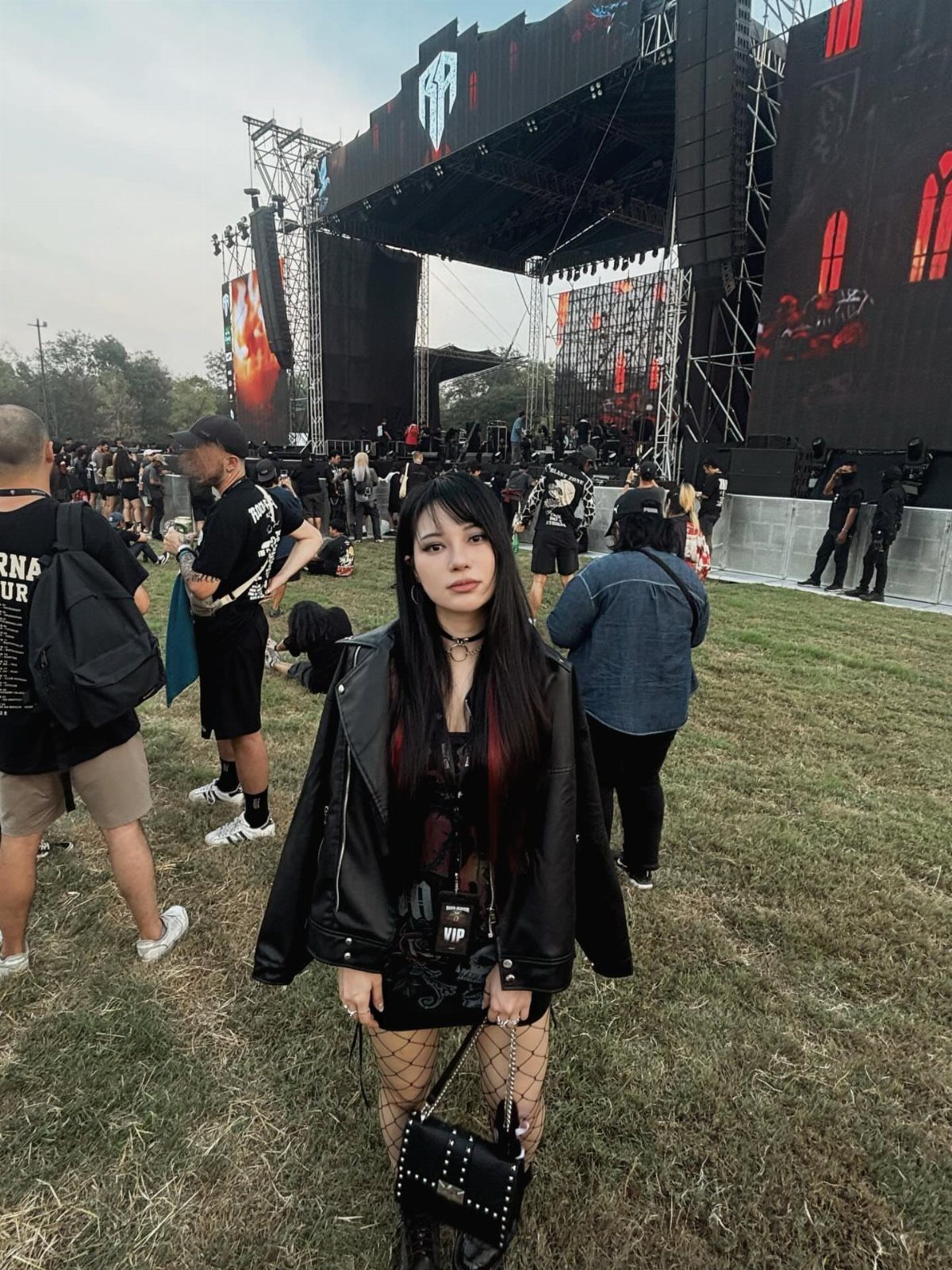 A woman in a black leather jacket and fishnets stands in a grassy outdoor concert field.