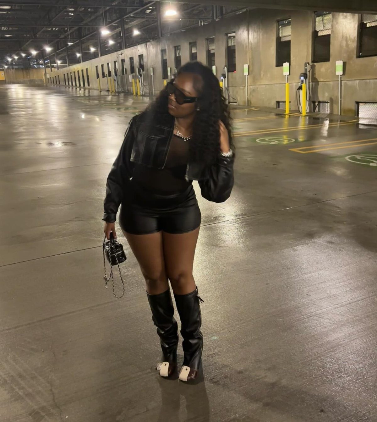 A woman wearing a black leather jacket, shorts, and tall boots standing on wet pavement.