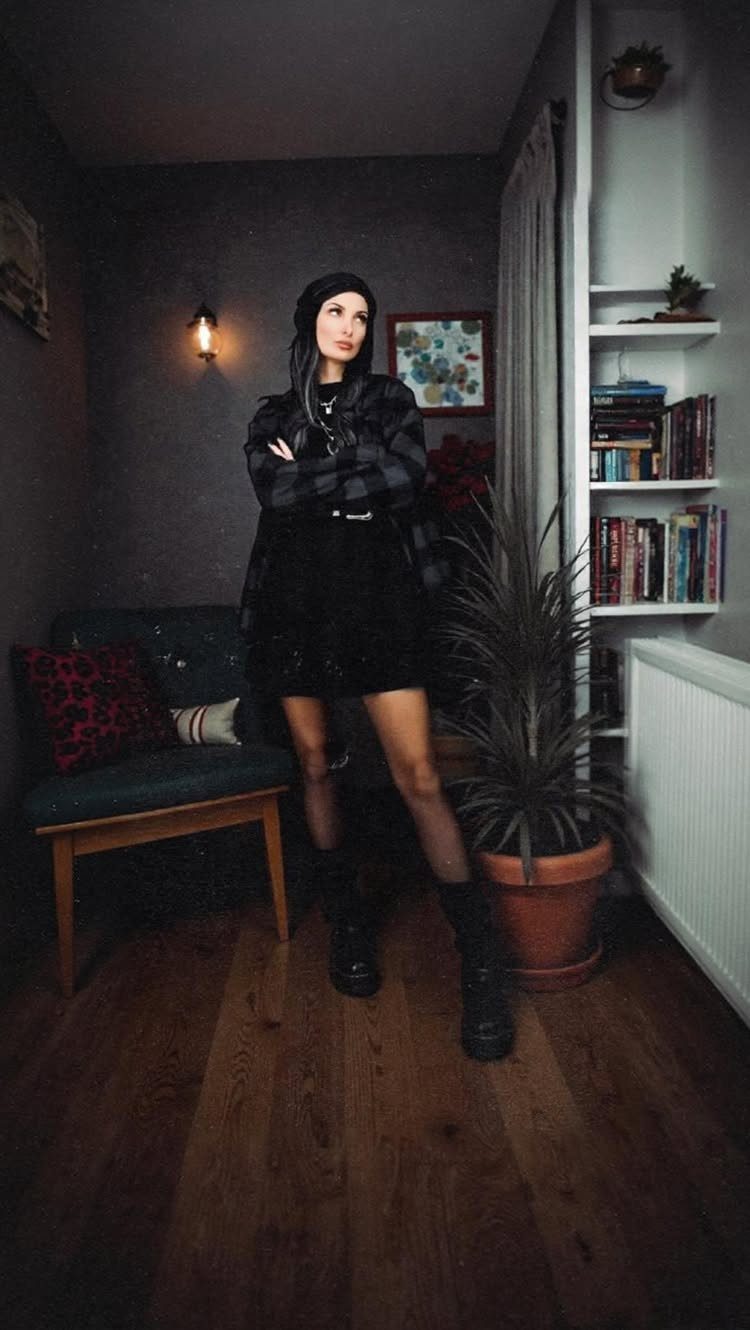 A woman posing in a black plaid flannel shirt and black combat boots indoors.