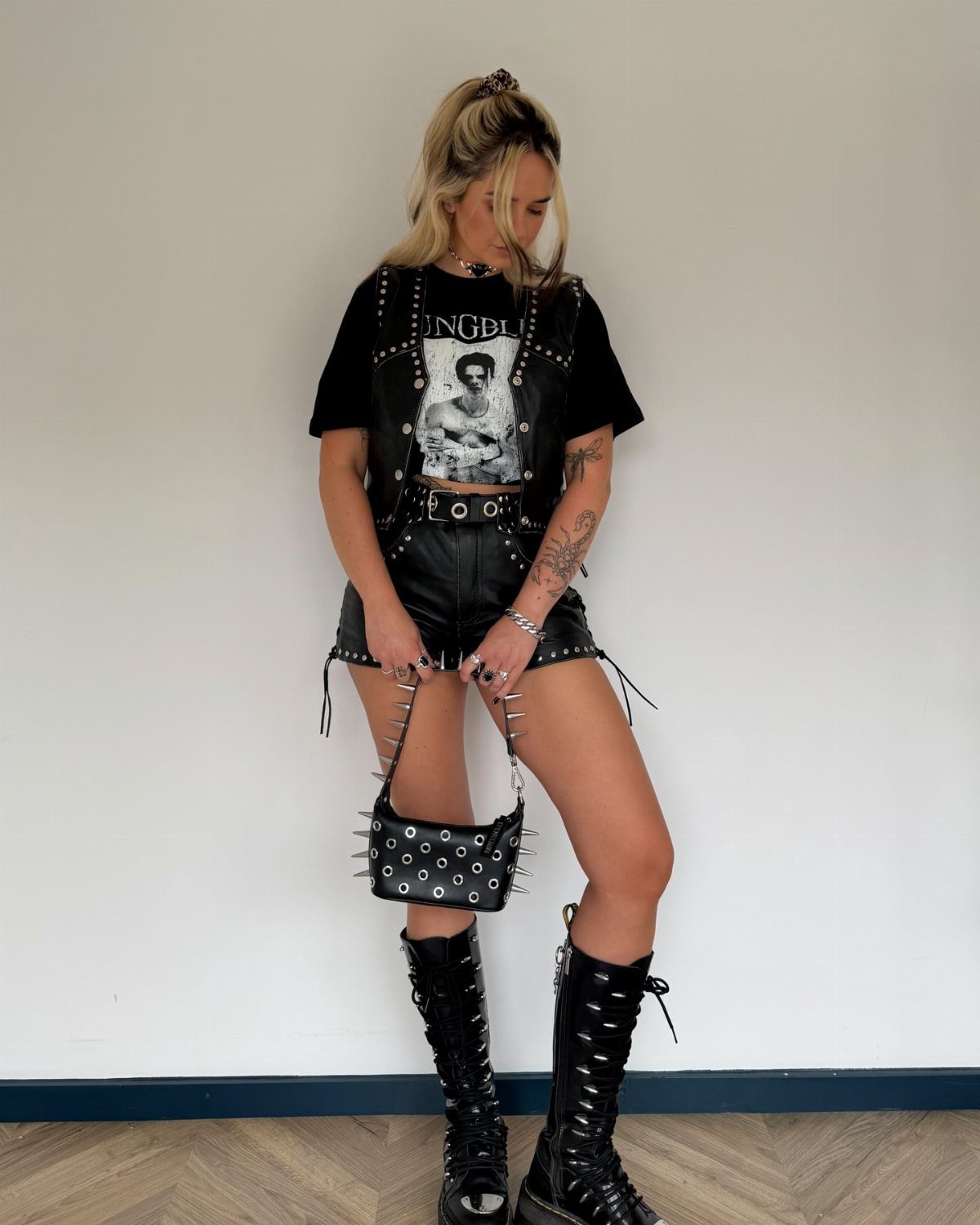 A woman wearing a black graphic tee, leather vest, leather shorts, and tall combat boots.