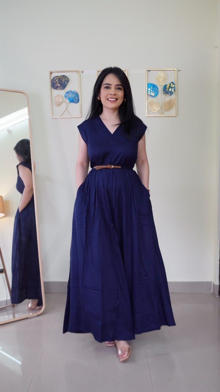 A woman smiling while wearing a long navy blue sleeveless maxi dress with a brown belt.