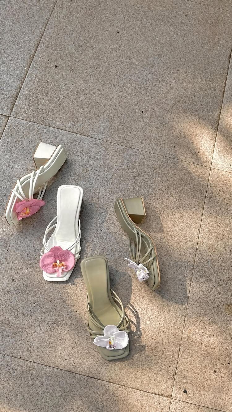 Two pairs of strappy sandals with floral decorations and block heels on stone pavement.