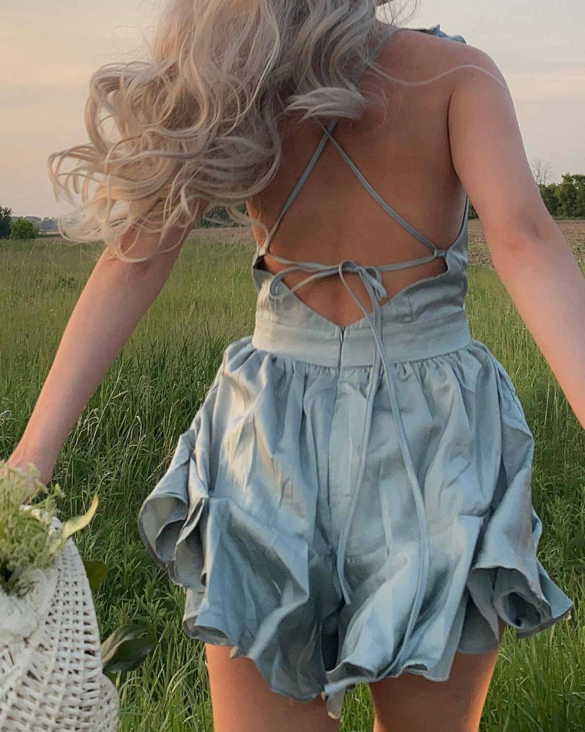 Woman in a light blue satin romper standing in a grassy field at sunset.