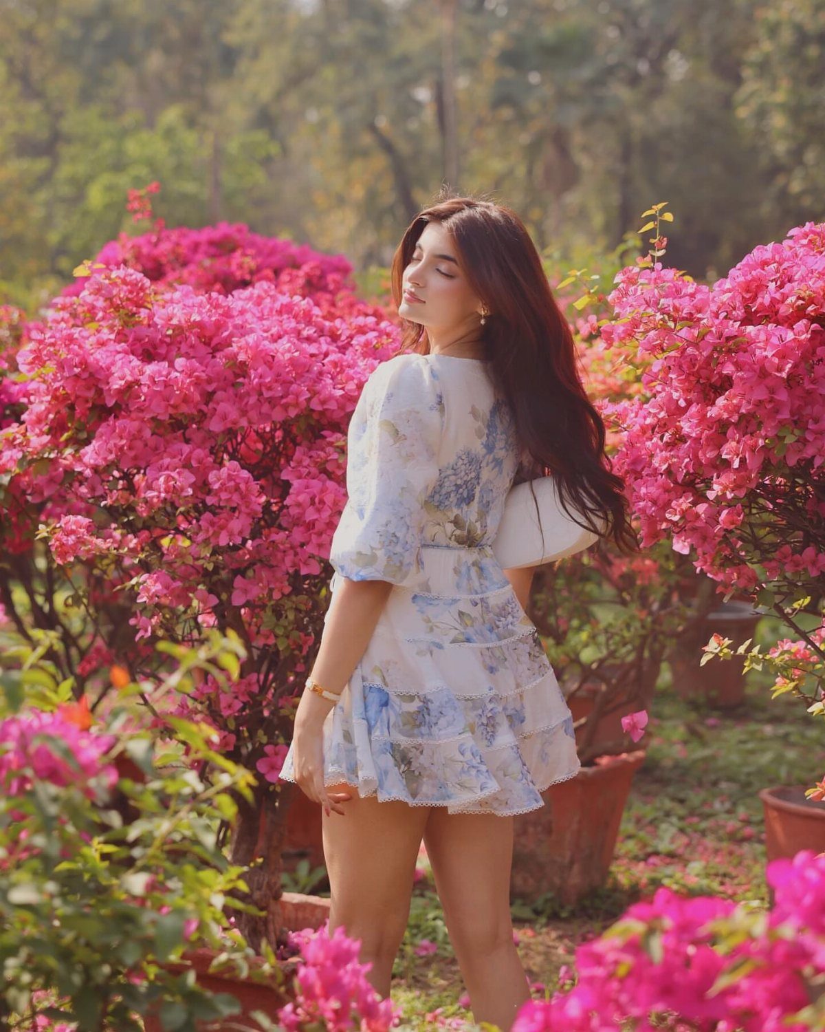 A woman in a white floral sundress standing amidst bright pink bougainvillea flowers in a garden.
