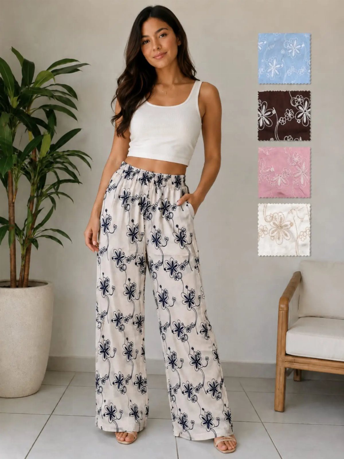 A woman wearing a white tank top and floral wide-leg trousers standing indoors.