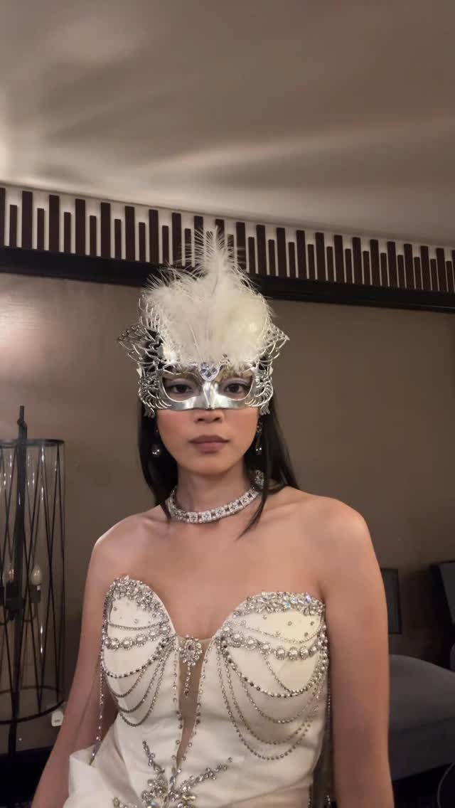 A young woman wearing a silver feathered masquerade mask and a beaded white gown.