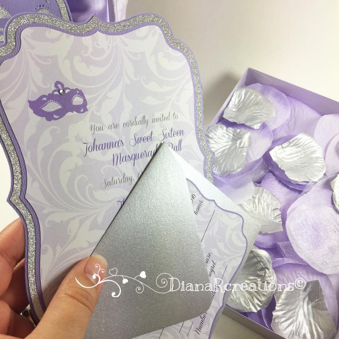 A hand holding a lavender and silver masquerade themed Sweet Sixteen invitation next to a box of petals.