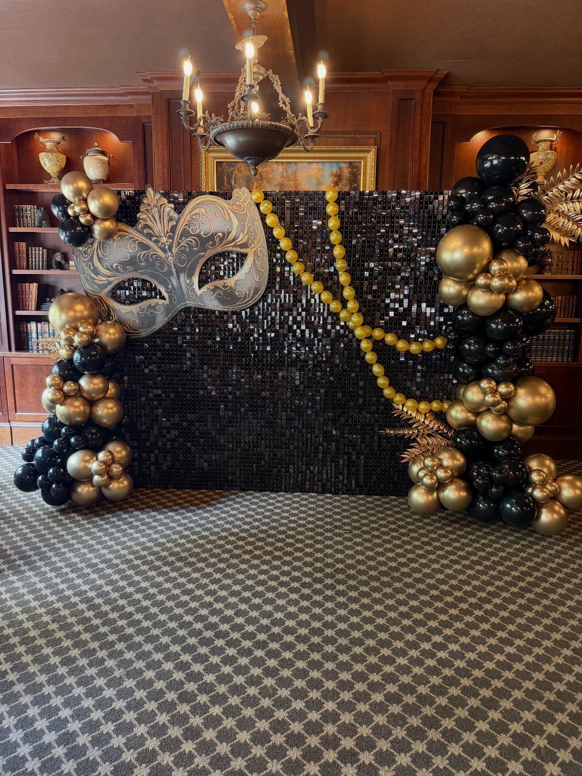 A black sequined backdrop featuring a silver masquerade mask and black and gold balloon decorations.