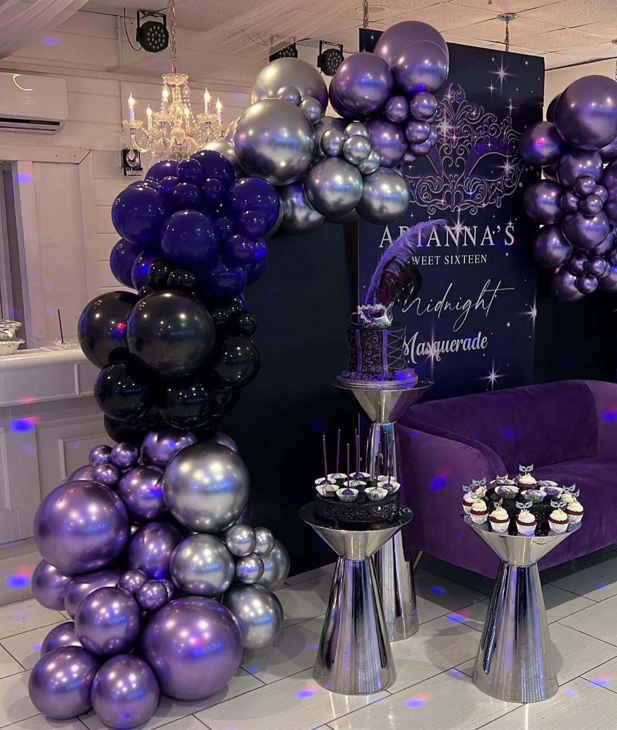 Purple and silver balloon arch surrounding a themed sweet sixteen masquerade party backdrop and dessert table.
