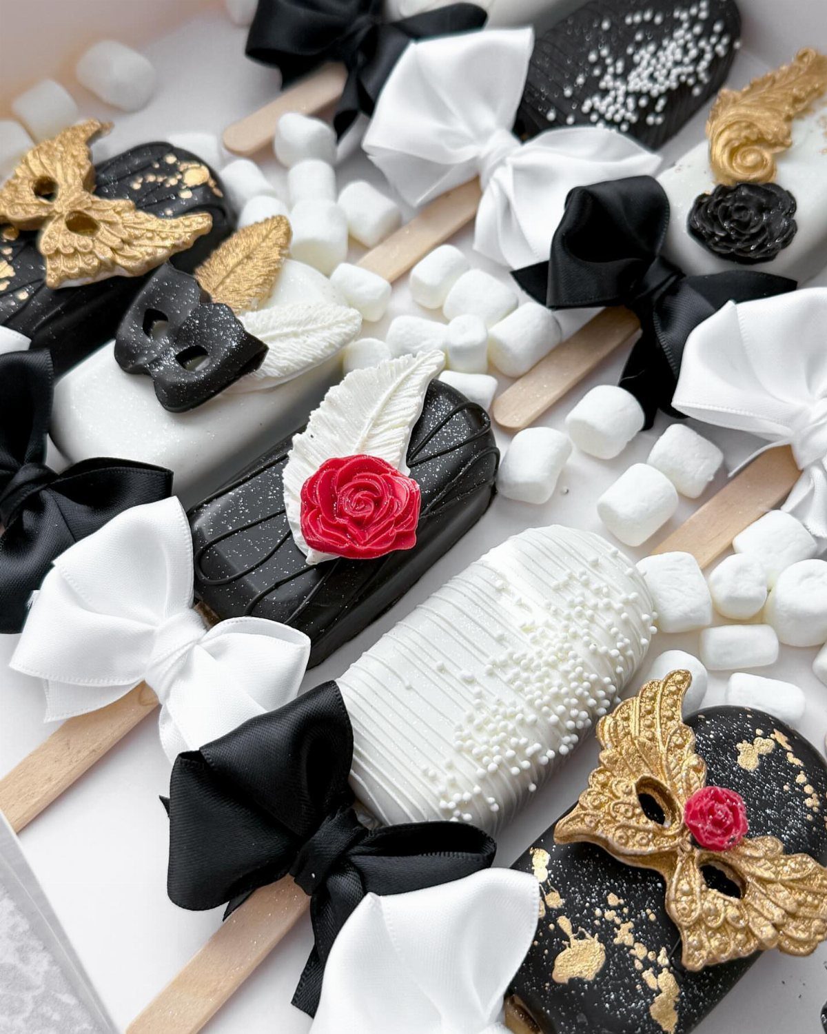 Decorated black and white cake pops with edible masks, roses, and satin bows on marshmallows.