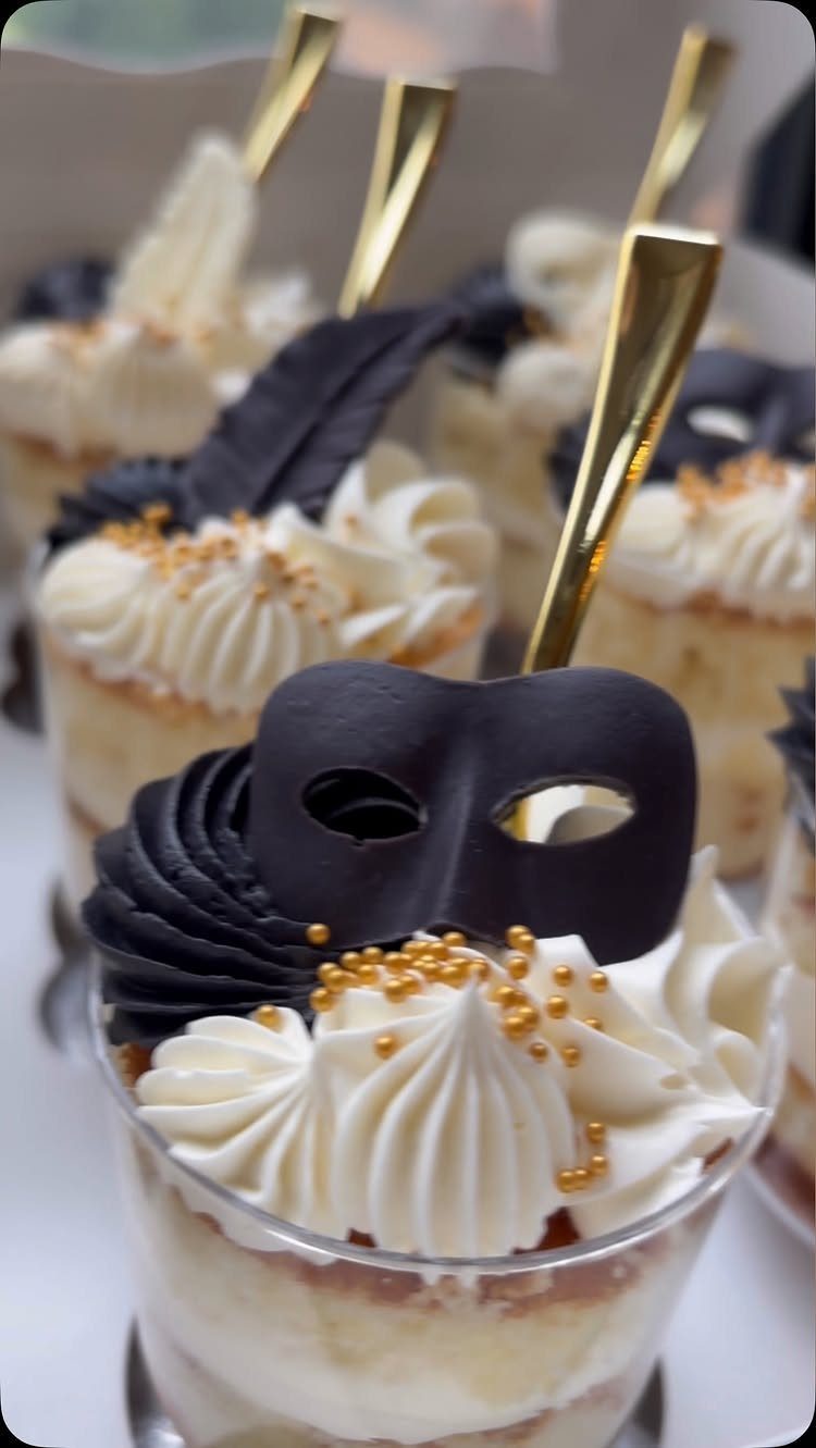 Dessert cups topped with white frosting, gold sprinkles, and a black chocolate masquerade mask.