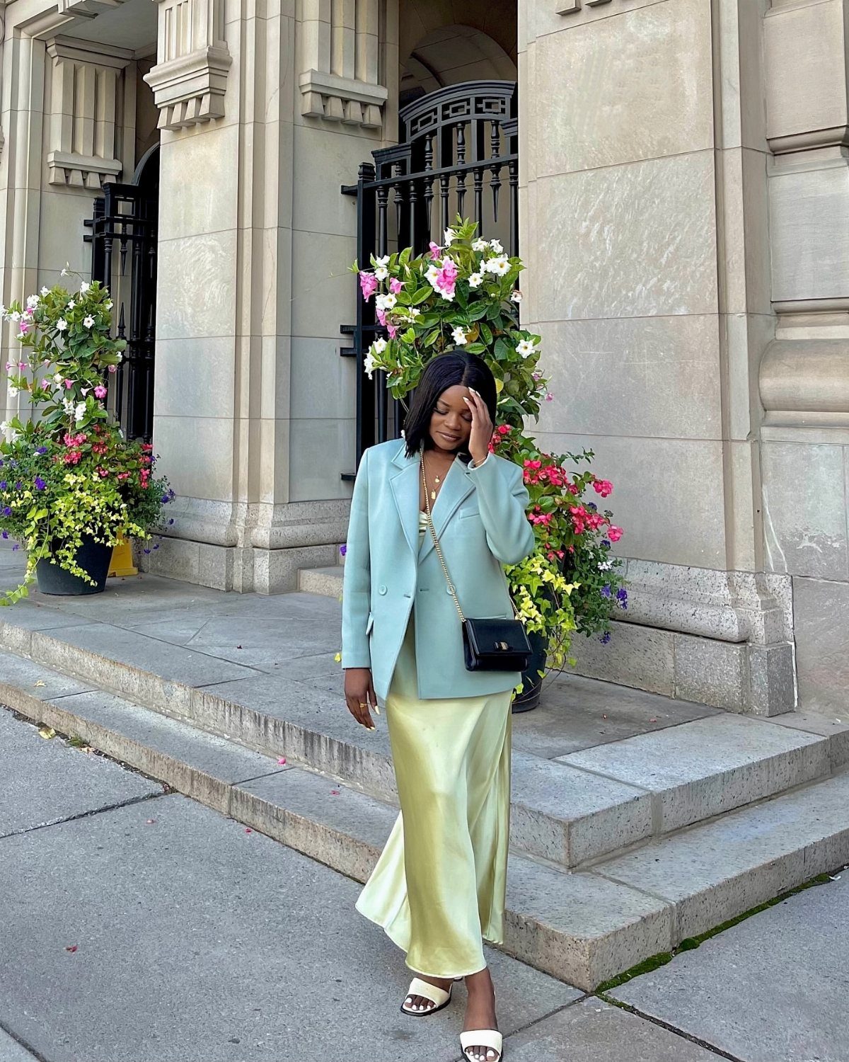 Woman in a light blue blazer and long yellow satin skirt standing near stone architecture.