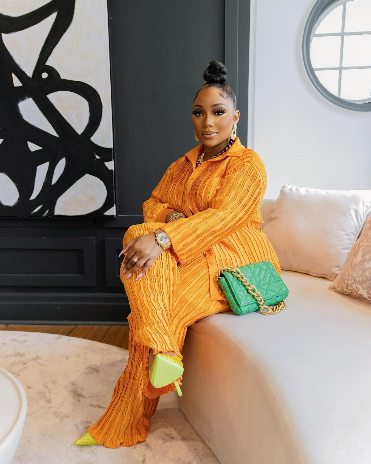 Woman in a bright orange textured outfit sitting on a white sofa with green accessories.