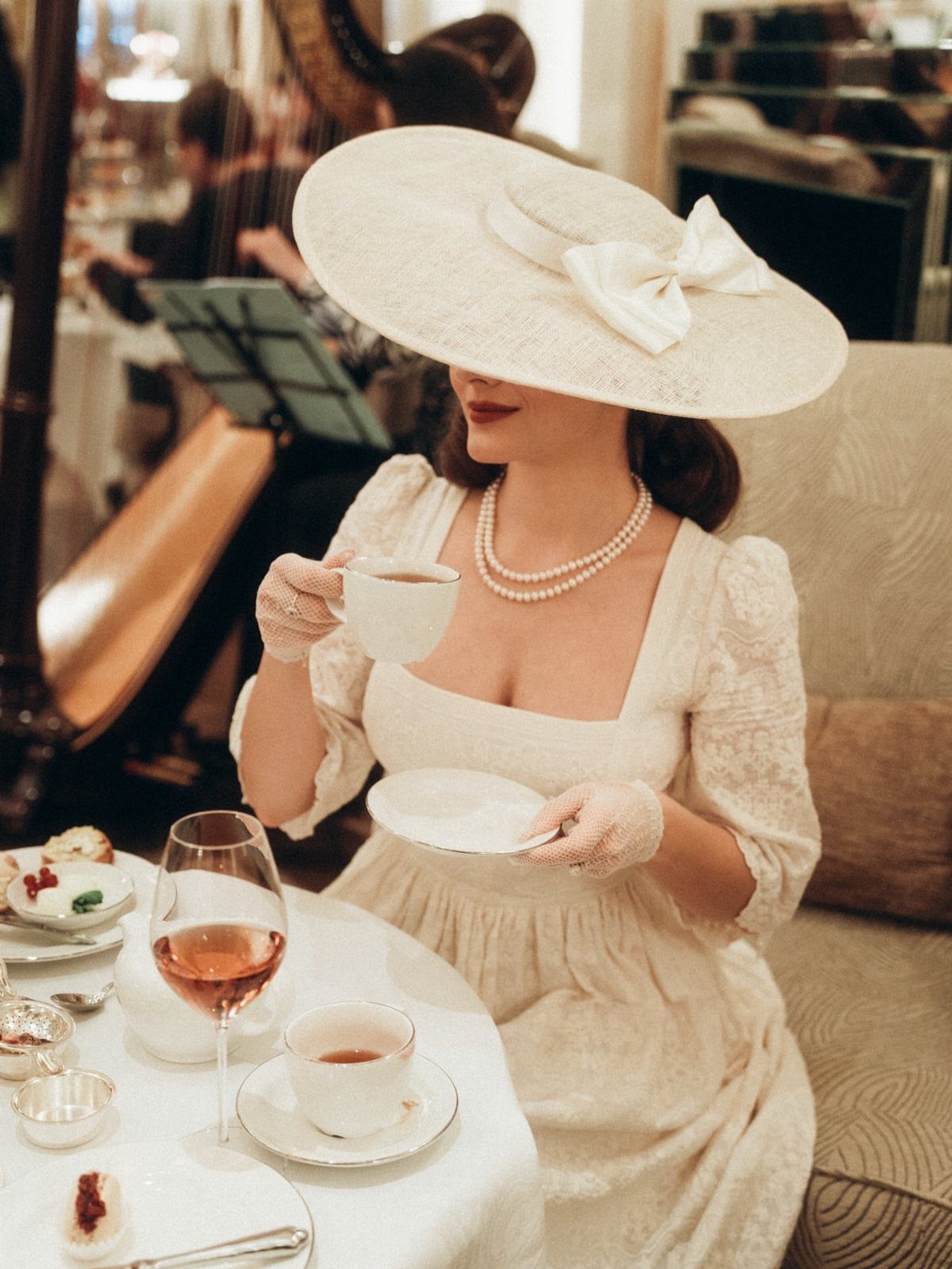 A woman wearing a large white wide-brimmed hat and lace dress enjoys tea at a table.