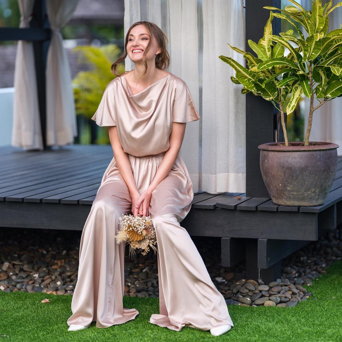 A smiling woman sitting on a wooden deck wearing a champagne satin wide-leg jumpsuit.