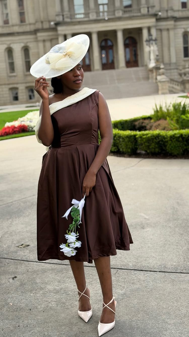 A woman in a brown tea-length dress and large white hat posing outdoors.