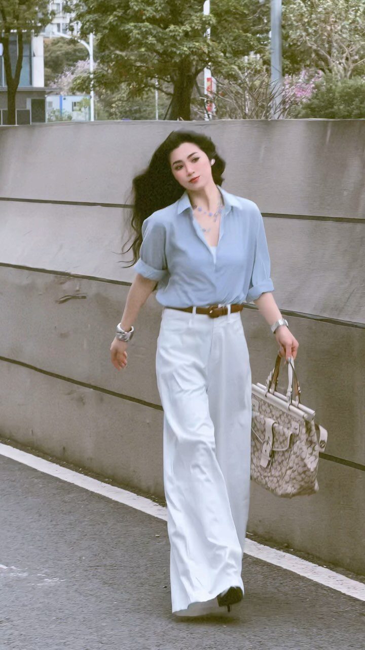 A woman walking in a light blue shirt and white wide-leg trousers carrying a handbag.