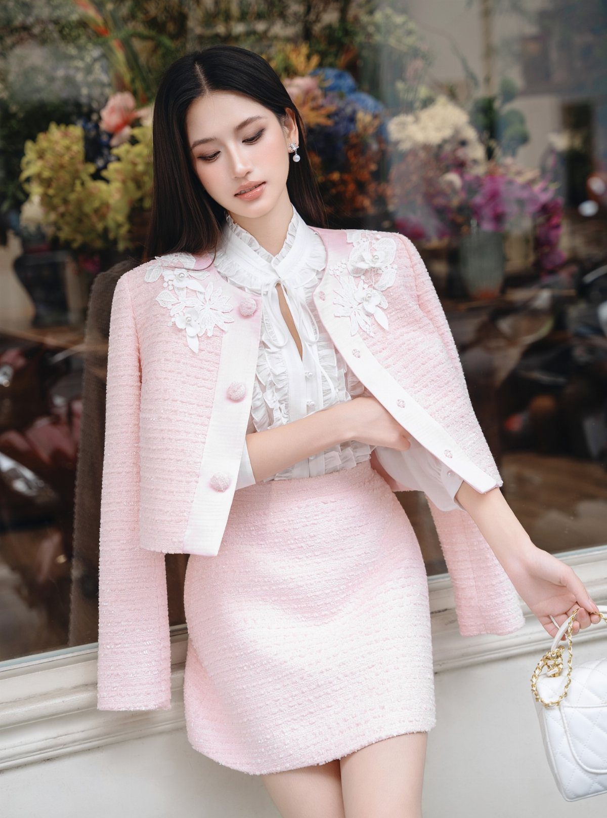 Woman wearing a pink tweed skirt suit and white lace blouse standing near flowers.