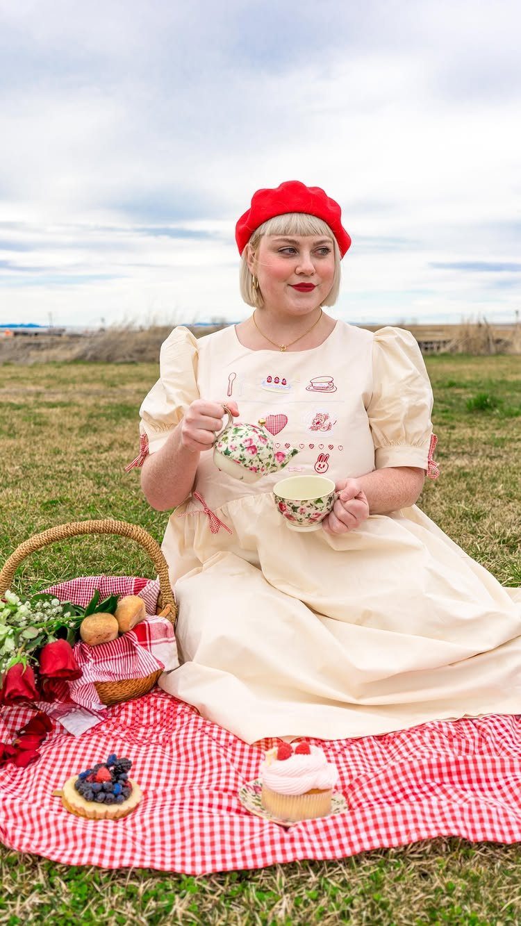 Woman in a cream puff-sleeve dress pouring tea during an outdoor picnic in a field.