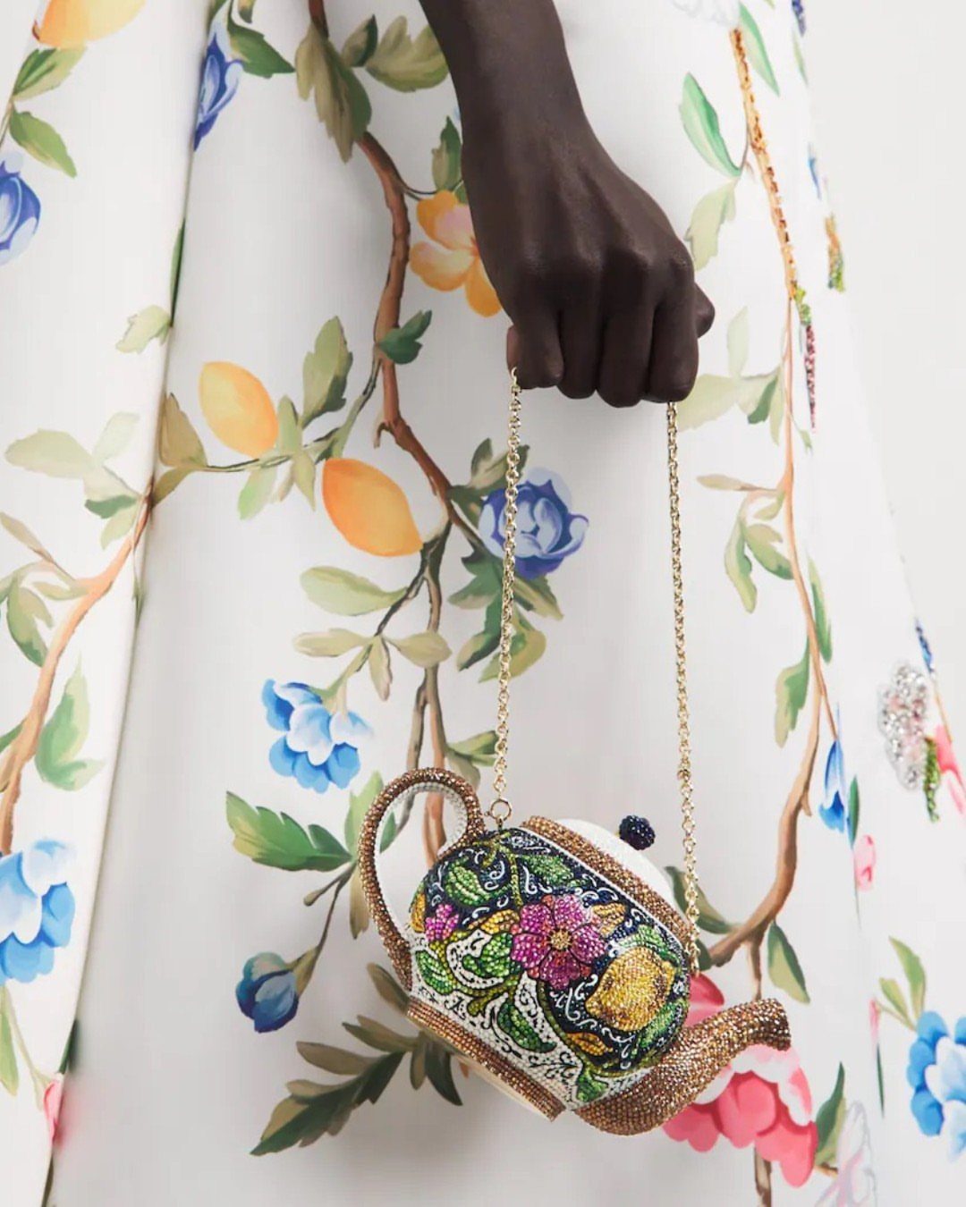 A hand holding a small, teapot-shaped beaded clutch with colorful floral embroidery and a gold chain.