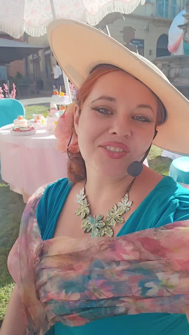 A woman wearing a wide-brimmed hat, teal dress, and floral shawl at an outdoor tea party.