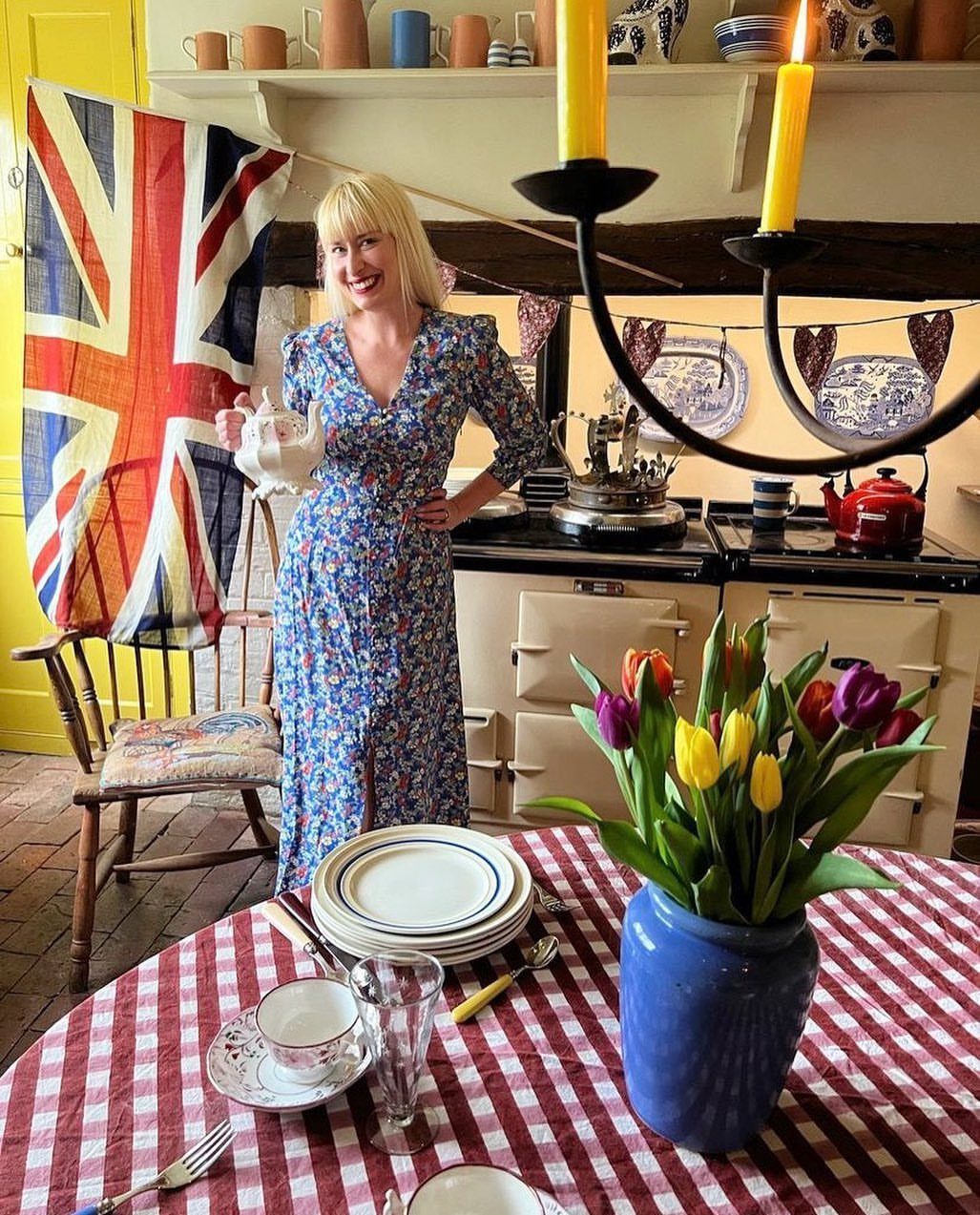 A woman in a blue floral dress holding a teapot near a set tea table.
