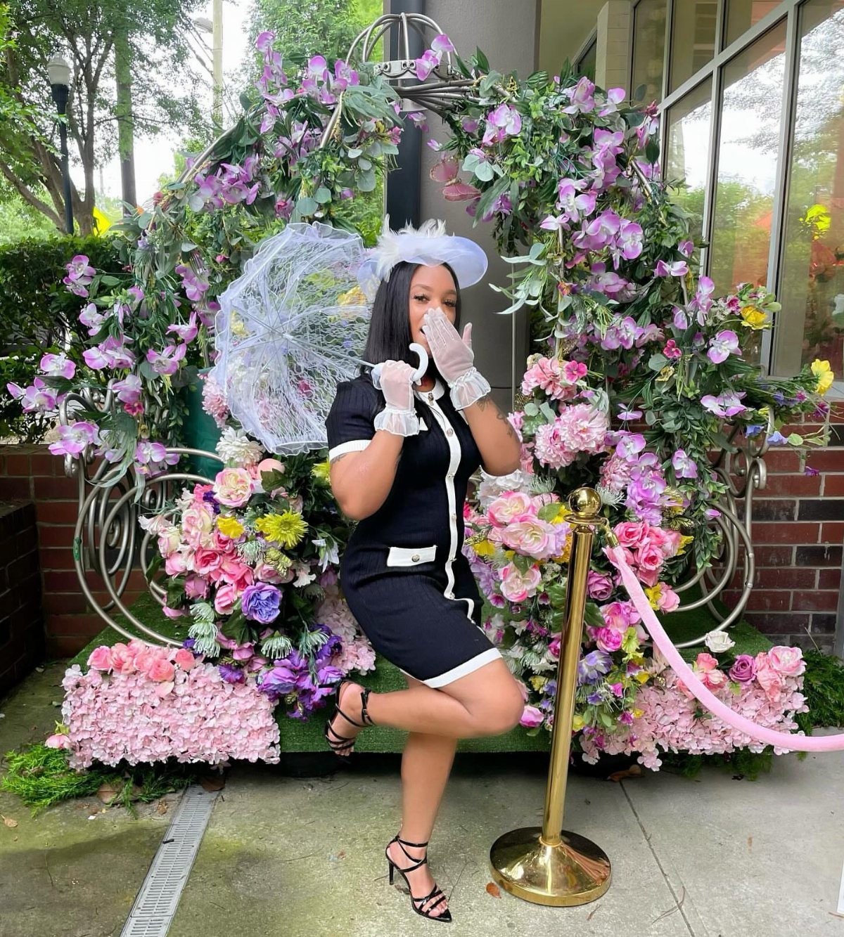 A woman wearing a white fascinator hat and gloves posing before a large floral arch.