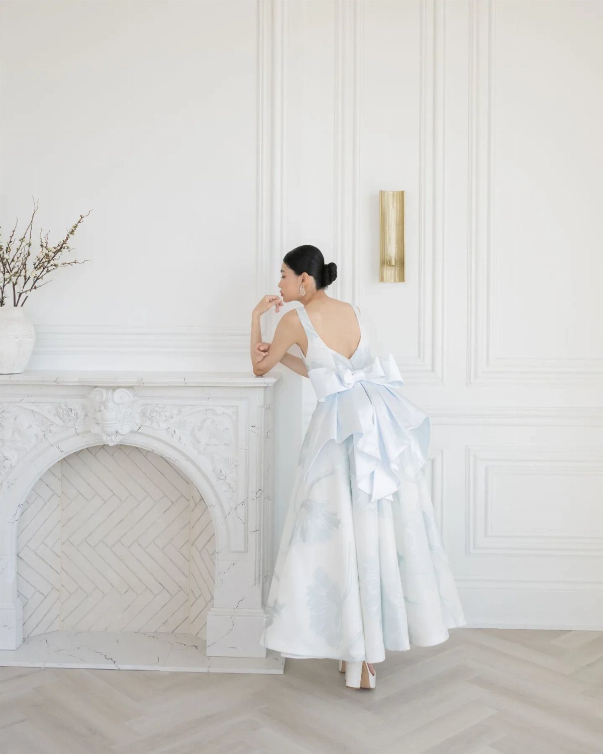 A woman in a light blue patterned gown with a large bow stands by a white fireplace.