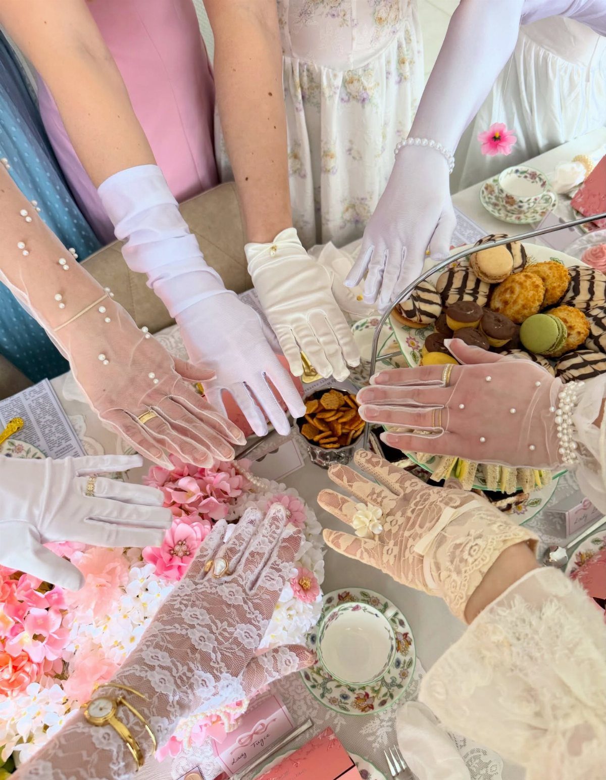 Hands wearing elegant lace and satin gloves reaching toward tea and pastries on a decorated table.