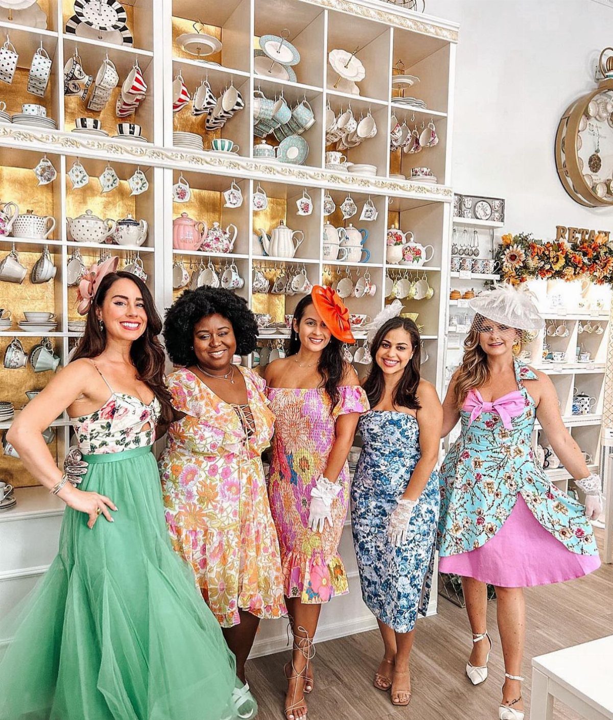 Five women smiling while wearing colorful pastel floral dresses in a tea shop.