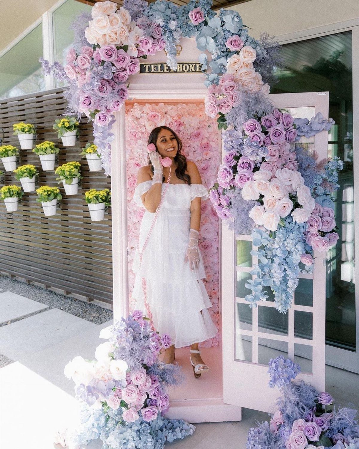 Woman in white dress posing inside a pink telephone booth adorned with pastel flowers.