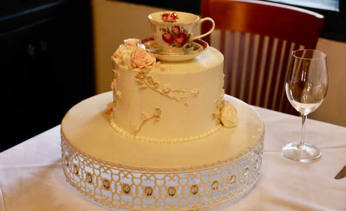 A white decorated cake topped with a floral teacup and pink roses on a white stand.