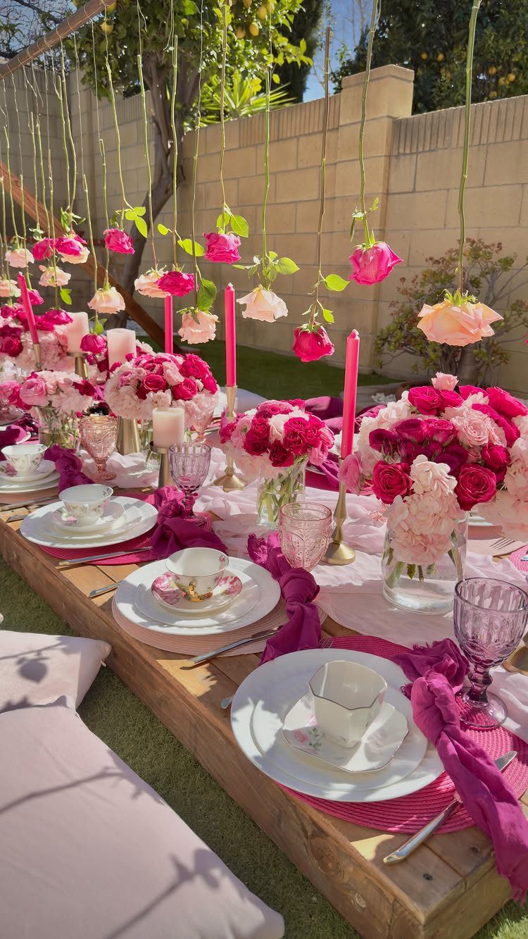 Pink and white floral arrangements decorate a low wooden table set for an outdoor tea party.