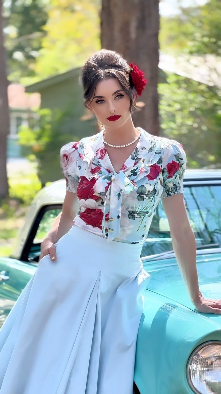 Woman in a floral blouse and light blue skirt posing by a vintage teal car.