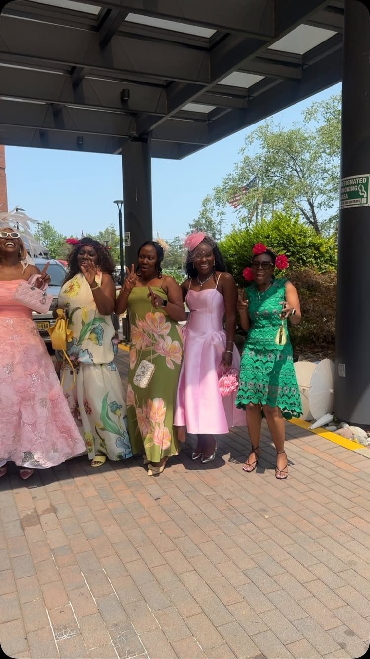 Five women posing outdoors in colorful, stylish dresses and decorative headpieces.