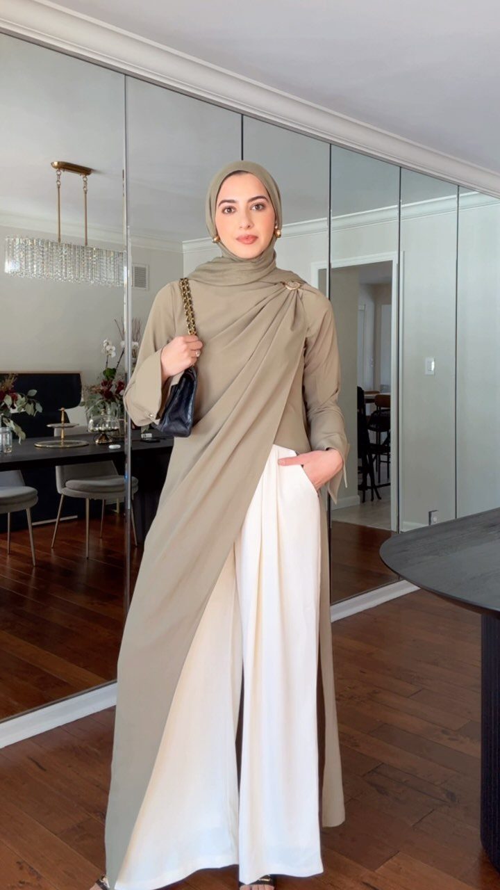 Woman in tan hijab and wrap top with wide-leg white trousers standing in a room.