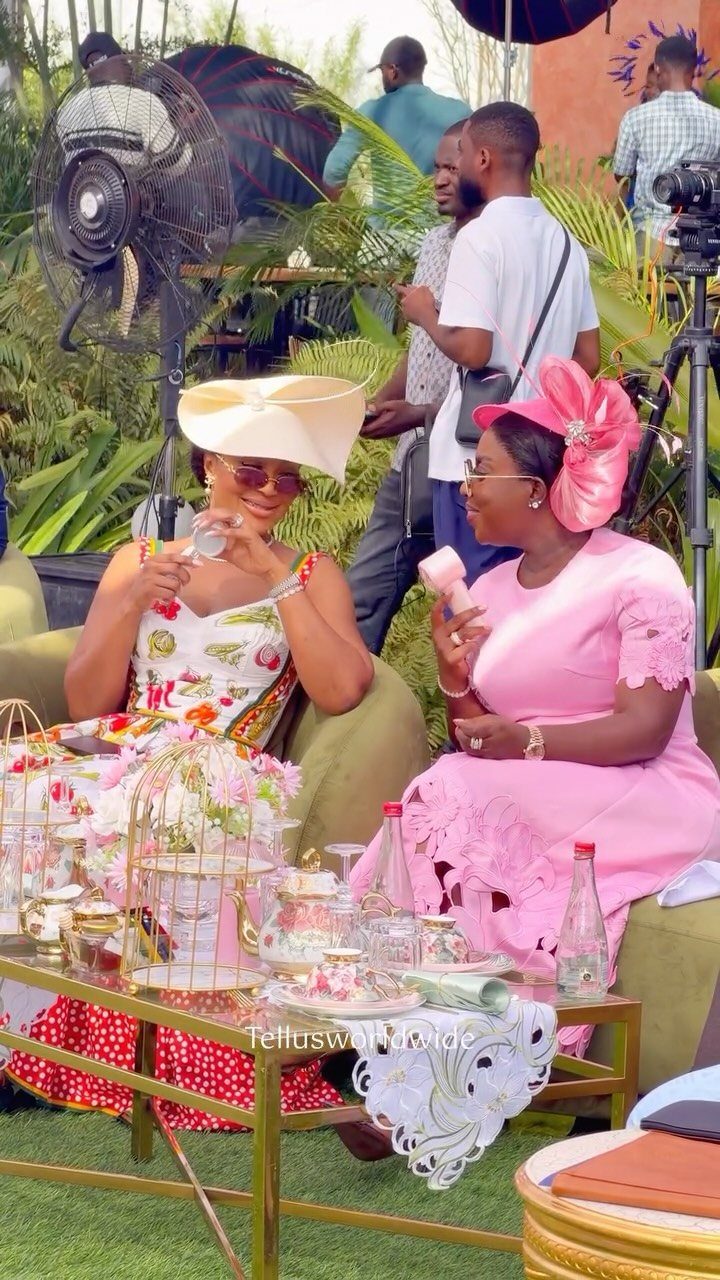 Two women in elegant dresses and hats enjoying tea at an outdoor garden party.