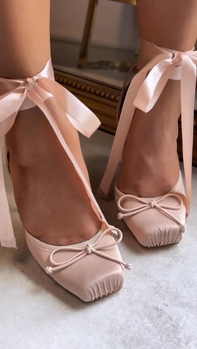 Pink satin ballet flats with long ribbons tied around the ankles on a light floor.