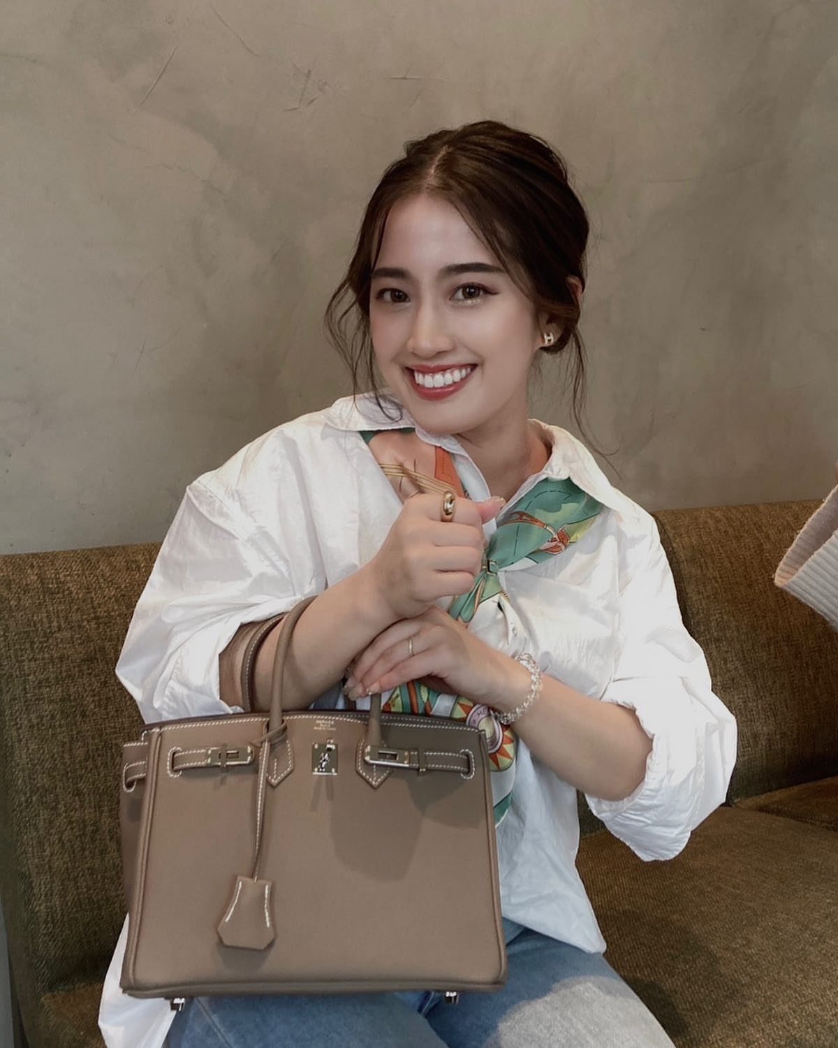 A smiling woman in a white shirt holding a large, structured beige leather handbag.