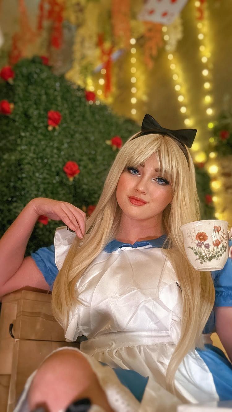 Blonde woman in Alice in Wonderland costume holding a floral teacup against a garden background.