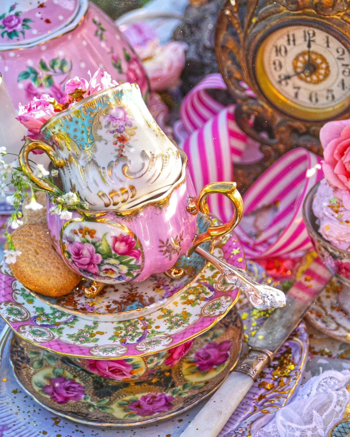 Mismatched floral teacups and saucers stacked with a vintage clock and pink ribbons.