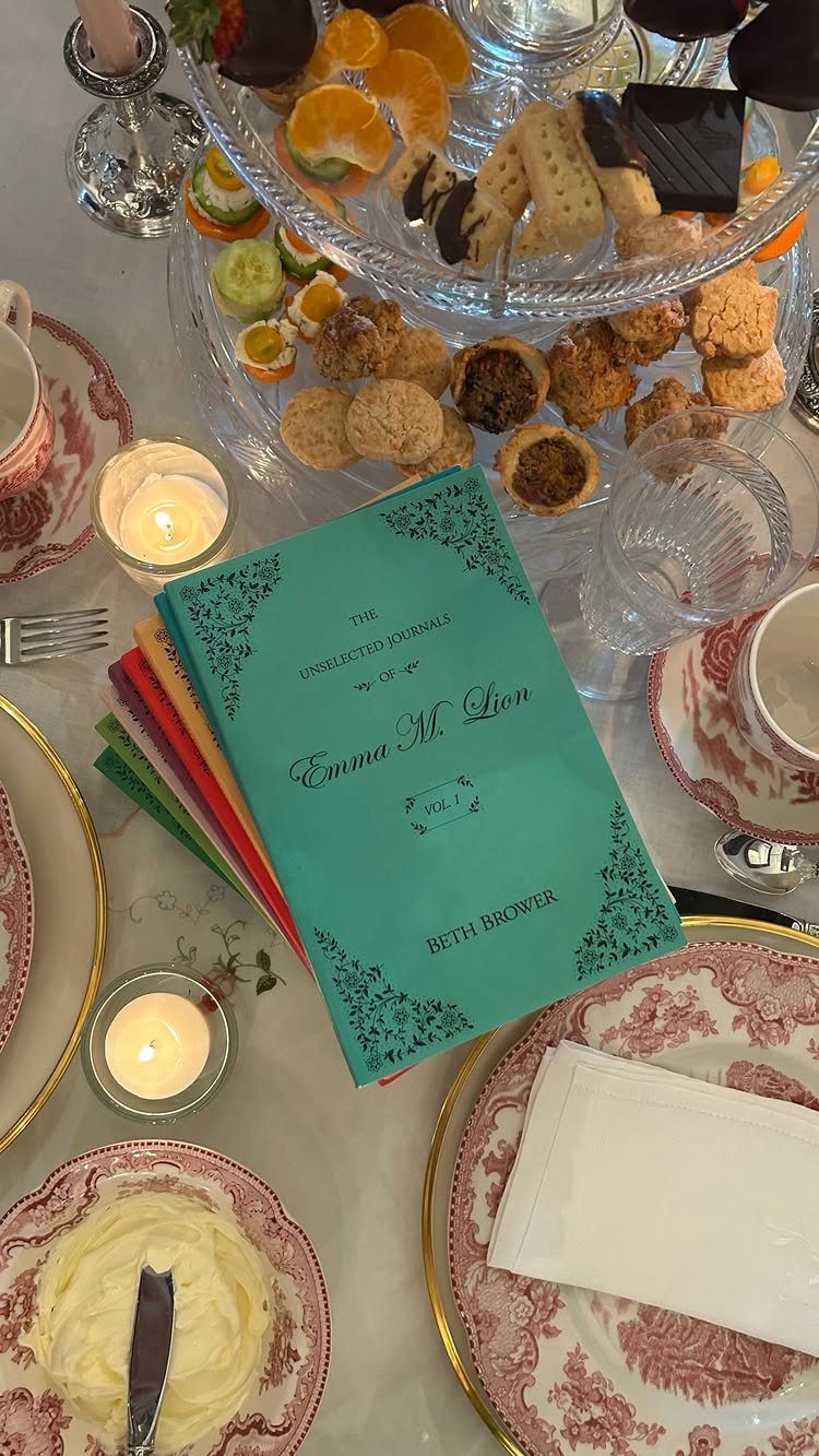 A teal book titled Emma rests among assorted pastries and lit candles on a tea party table.
