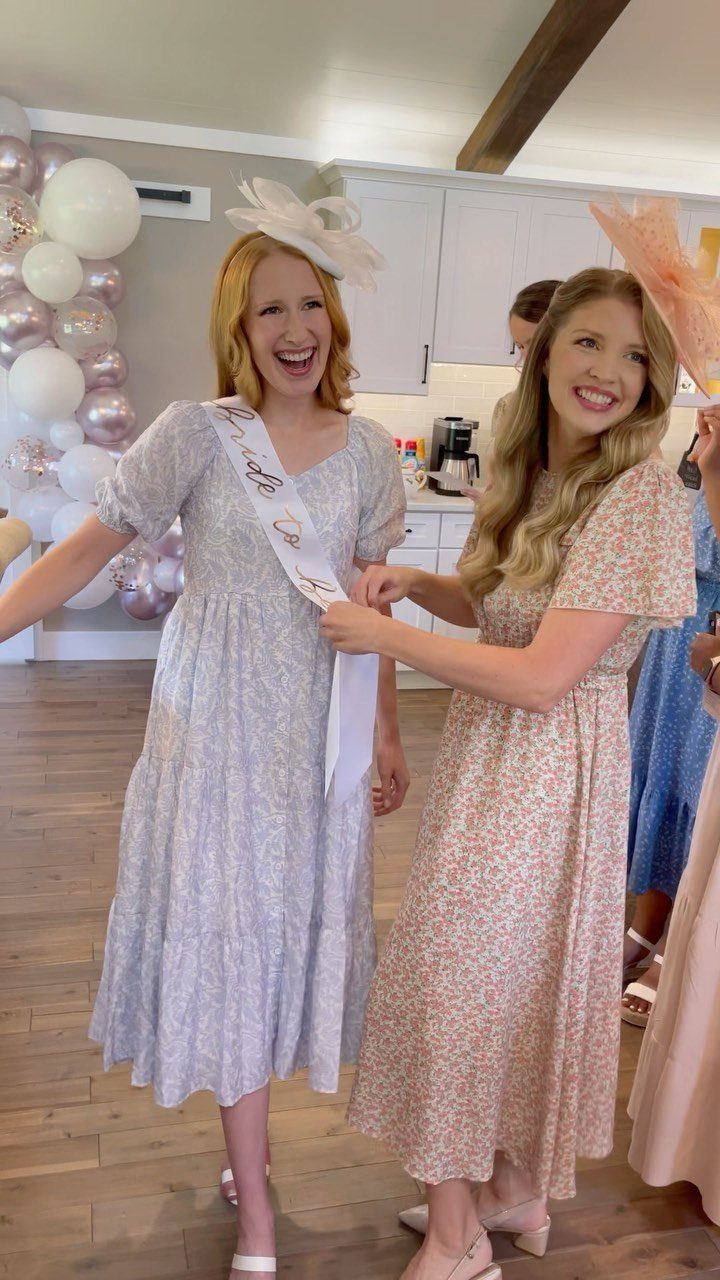 Two women in floral dresses smiling while one adjusts a white sash on the other.