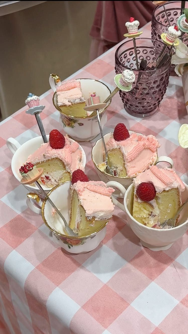 Pink frosted cake slices with raspberries served inside decorative floral teacups on a checkered tablecloth.