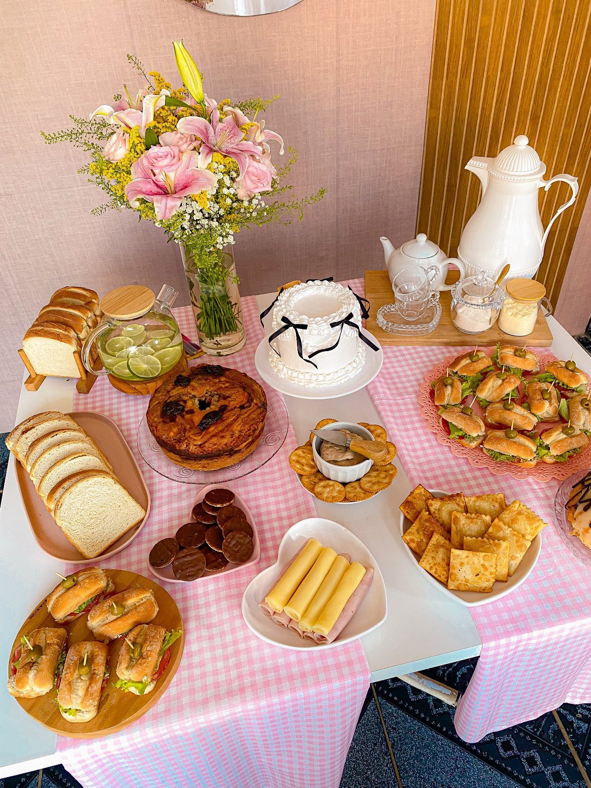 A pink checkered table spread with sandwiches, cake, pastries, and a large bouquet of pink lilies.