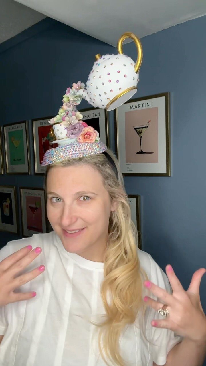 Woman wearing a large, decorative teacup-shaped fascinator hat adorned with flowers and rhinestones.