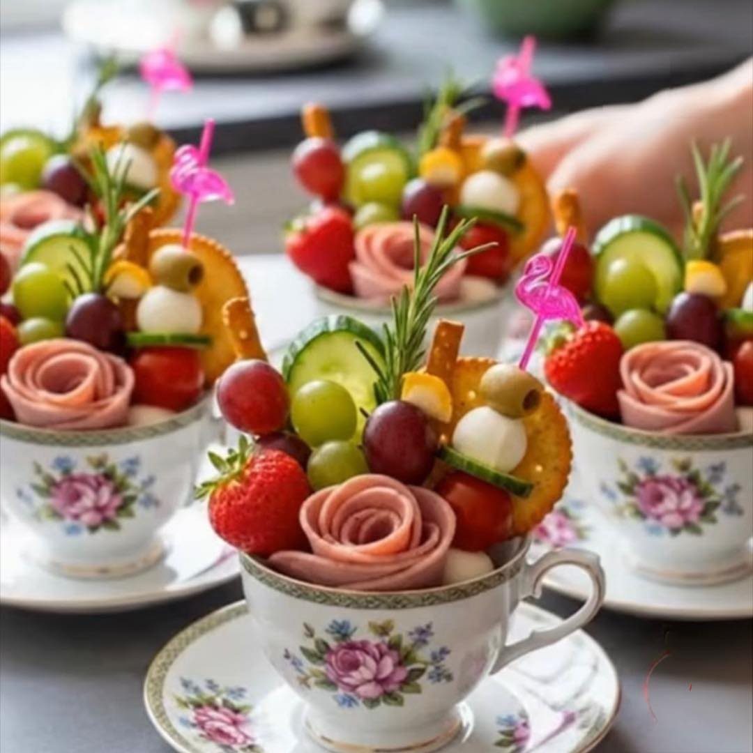 Savory snacks like ham roses and grapes served inside floral patterned teacups.