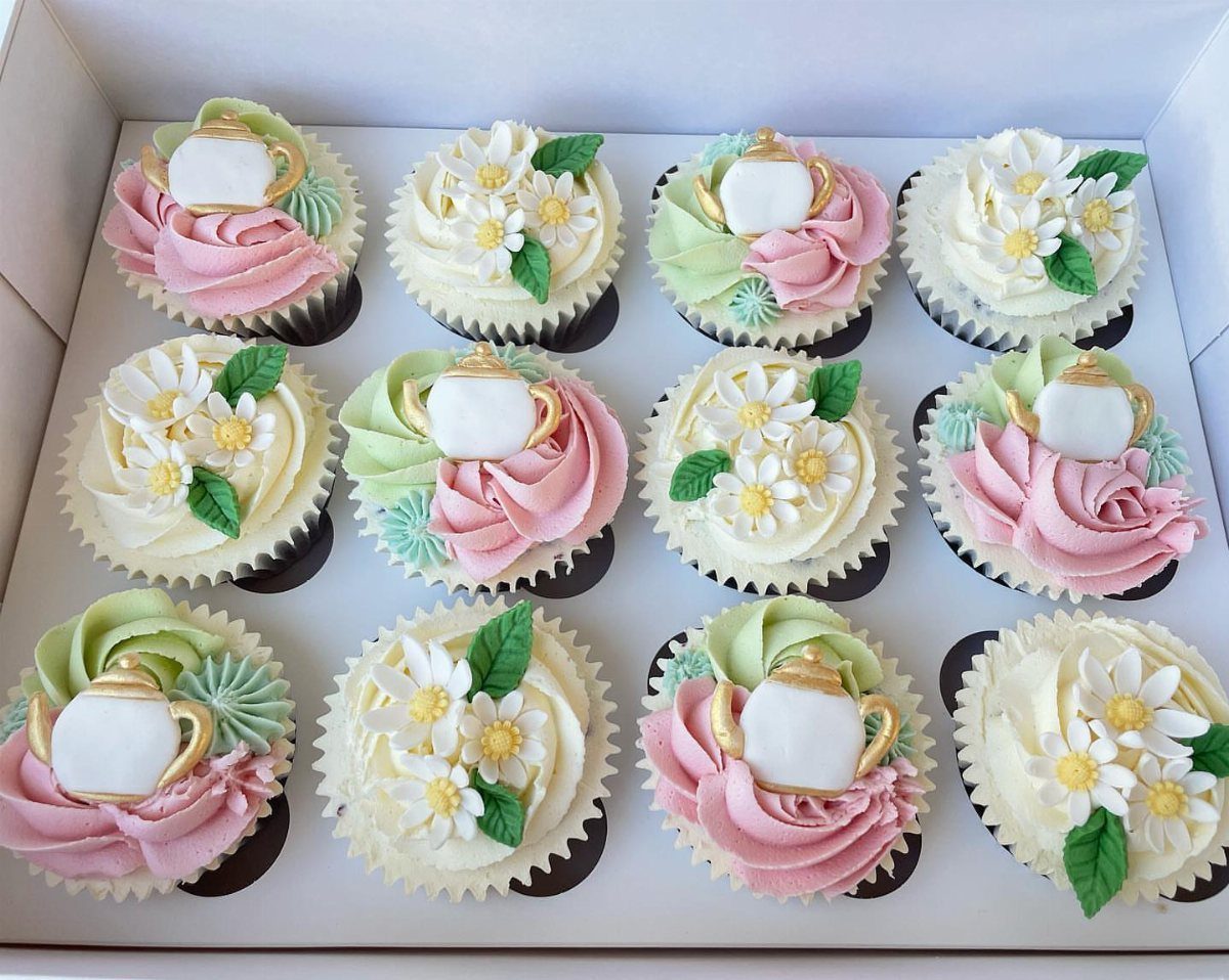 Twelve decorated cupcakes featuring teapot, rose, and daisy frosting designs in a white box.